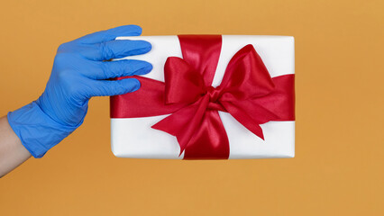 Christmas present. Gifts surprise. Delivery hygiene. White congratulation box with red ribbon in hand in protective gloves isolated on yellow background.