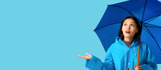 Beautiful young Asian woman in raincoat holding umbrella on light blue background with space for text - obrazy, fototapety, plakaty