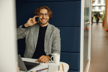 Smiling male boss talking phone with client while sitting in coworking space and working on laptop