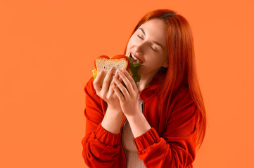 Young redhead woman eating tasty sandwich on orange background