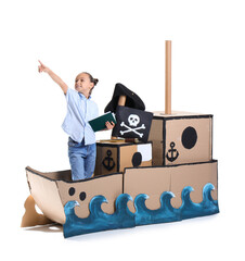 Little girl with adventure book pointing at something in cardboard pirate ship on white background