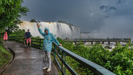 A man is standing on the path leading to the waterfall, leaning on the railing. Hand raised, smiling. In the distance, you can see the pedestrian observation bridge for tourists. Iguazu Falls. Brazil. - Powered by Adobe