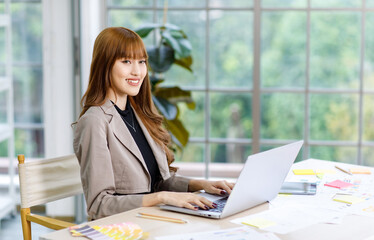 Asian professional successful young female businesswoman creative graphic designer employee in casual fashionable suit blazer outfit sitting working using typing laptop computer at office workstation