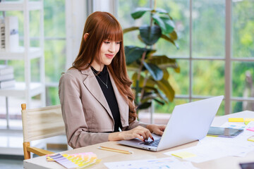 Asian professional successful young female businesswoman creative graphic designer employee in casual fashionable suit blazer outfit sitting working using typing laptop computer at office workstation