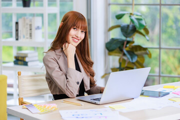 Asian professional successful young female businesswoman creative graphic designer employee in casual fashionable suit blazer outfit sitting working using typing laptop computer at office workstation