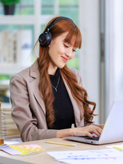 Asian professional successful young female businesswoman creative graphic designer in casual outfit sitting smiling wearing headphone listening to music playlist while working typing laptop computer