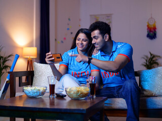 Indian man and a woman watching a live cricket match at home - Cricket Cup match  1-day match. Couple wearing Indian jersey - watching sports  cricket fever  T20 series  happy gesture