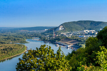 Fototapeta premium A plant or factory on the banks of the Volga near the city of Samara, Russia against the backdrop of the Zhiguli mountains. Blue water, tree crowns, forest. Quiet summer morning with light sky.