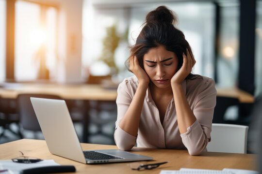 Exhausted businesswoman having a headache in modern office. Mature creative woman working at office desk with hands on head feeling tired. Stressed casual business woman feeling eye pain.