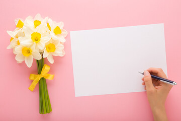 Woman hand holding pen and writing on greeting card. Fresh white narcissus bouquet with yellow ribbon on pink table background. Pastel color. Empty place for text, quote or sayings. Top down view.