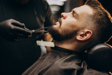 Handsome man cutting beard at a barber shop salon - obrazy, fototapety, plakaty