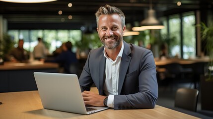Happy smiling middle aged professional business man company executive ceo manager wearing blue suit sitting at desk in office working on laptop computer laughing at workplace. Portrait.