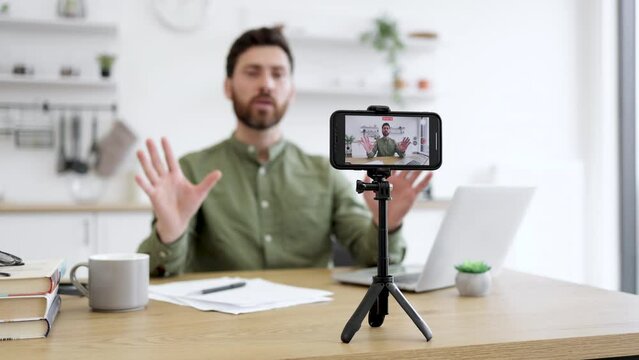 Blurred background of male streamer meditating with closed eyes and holding mudra gesture while filming online course at home. Focus on wireless smartphone fixed on tripod and recording video.