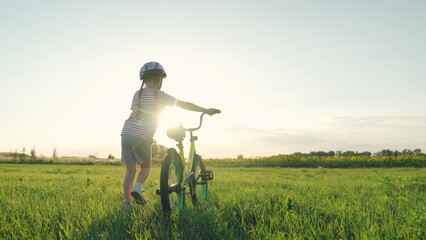 Cute little girl wearing helmet walks pulling bicycle across summer field