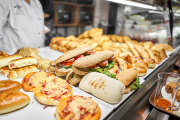 Fresh sandwiches, pies and pizza to choose from in the window of a cafe, buffet or self-service cafe. Croissant with ham and cheese. Hot pastries lie on the shelf in the cafe. Buns in the store. - 663151367