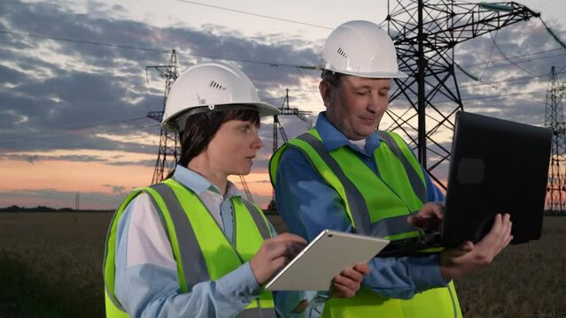 Engineers with colleague study data on computers by power transmission lines