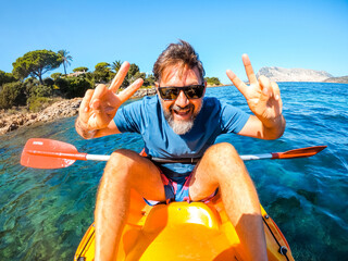One cheerful man have fun and pose for a crazy picture sitting inside a yellow kayak canoe  with ocean water and coast in background. Happy tourist summer holiday vacation lifestyle people doing tour - obrazy, fototapety, plakaty