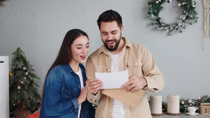 Happy young couple stand in the kitchen during the Christmas period, opening a letter reading pleasant, amazing, positive news. The man and woman received news of a mortgage approval or a lottery win.