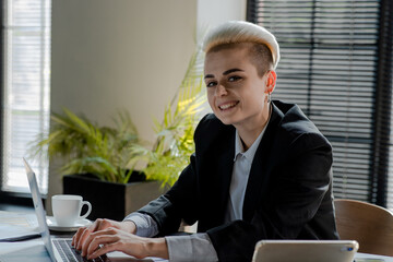 Young caucasian woman working in modern office using her laptop. Smiling businesswoman sits indoors at coworking room with computer. Happy girl project manager texting notebook to colleagues