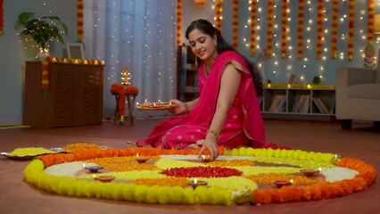 Happy young Indian woman placing diya lamps on decorated flower rangoli for diwali festival celebration at home - concept of festive preparation, traditional culture and spirituality