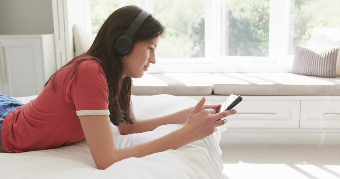 Happy biracial woman in headphones lying on bed in sunny room using tablet, copy space, slow motion