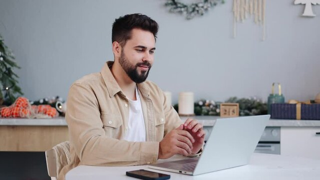 Handsome man in a beige shirt and white tee sits in a festively decorated kitchen during Christmas, making online orders or purchases gifts with a laptop, then looks up with a smile at the camera.