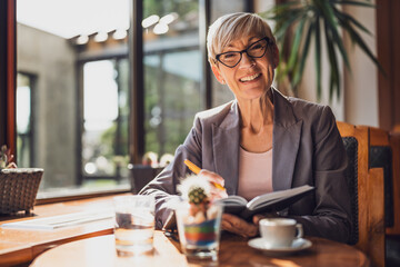 Mature woman is sitting in cafe and relaxing. She is drinking coffee and using note pad.