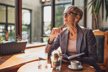 Mature woman is sitting in cafe and relaxing. She is drinking coffee and using note pad.