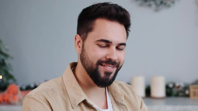 Positive man in a beige shirt and white tee sits in a festively decorated kitchen during Christmas, making online orders or purchases gifts with a laptop, then looks up with a smile at the camera.