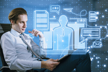 Attractive young european businessman with laptop and creative blue devices hologram sitting on blurry office interior background. Digital marketing, social network and online service concept.