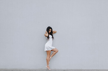 Photography, portrait of a stylish beautiful young brunette girl with curly hair, Armenian woman in a white dress in the city against the background of a wall outdoors.