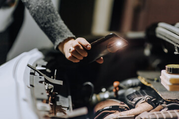 A woman mechanic repairs a car with a breakdown in the engine under the hood using tools at night, highlighting it with a flashlight on her phone or smartphone. Close-up photography, portrait.