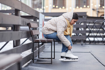 Side view portrait of young woman putting on skates in ice skating rink outdoors, copy space