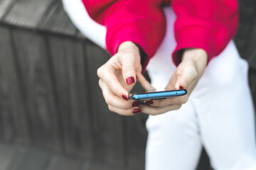 Close up Woman hands in red hoodie with red manicure is holding smartphone on bench outdoors.