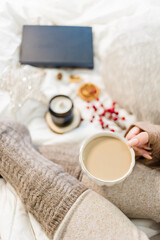 Cozy winter scene. Christmas vibes. Woolen socks. Woman having coffee in bed with book and candle.