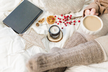 Cozy winter scene. Christmas vibes. Woolen socks. Woman having coffee in bed with book and candle.