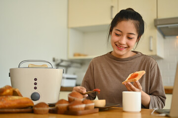 Smiling young woman spreading strawberry jam on slice of bread at kitchen table