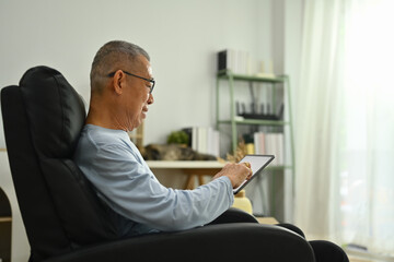 Side view of 70s elderly man sitting on the armchair and using digital tablet