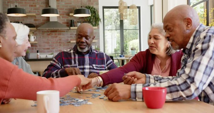 Happy diverse senior female and male friends talking and doing jigsaw puzzle in kitchen, slow motion