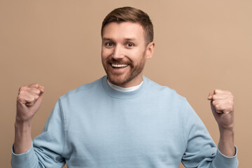 Fototapeta premium Happy cheerful man with crazy face expression showing win victory gesture on beige background looking at camera. Guy winner celebrates success, achieving goal. Body language, sincere emotions concept.