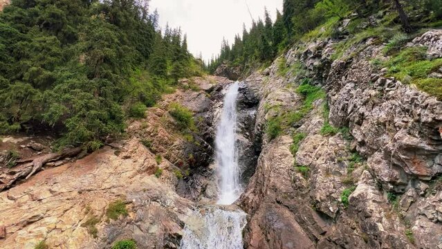 Waterfall of the Barskun gorge "Manas Bowl". A picturesque waterfall flows between dense spruce forests and rocks. Sun glare on the water. Splashes of water. Issyk-Kul region, Kyrgyzstan. 4K