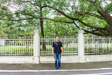 Asian Black T-Shirt is standing in front of white vintage fence beside the road in the afternoon time.