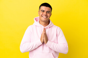 Young handsome man over isolated yellow background keeps palm together. Person asks for something