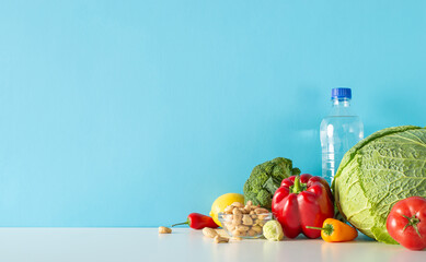 Balanced living illustration: side view table composition comprising water bottle, cashew nuts, medley of vegetables, like broccoli, pepper and more set against blue wall for text or promo content
