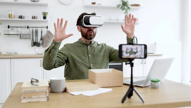 Excited caucaisan blogger testing VR headset while recording video on modern smartphone fixed on tripod. Bearded man making review of new gadget during unpacking of online purchase at home.