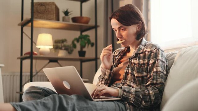 Portrait Of Pretty Young Woman Freelancer Having Distance Remote Work Typing Browsing Scrolling Products In Internet Online Store On Laptop Computer And Eating Cookie From Pocket At Light Home Indoors