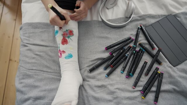 Hands teenage girl with broken leg in orthopedic cast painting pictures felt tips sitting in bed at home. Teen drawing on cast relaxing. Recovery damaged leg after injury, forced sick leave concept.