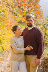 Fototapeta premium Beautiful couple in sweaters hugging on a background of yellow and red leaves in the autumn forest. Concept: vacation trip weekend in the forest autumn among trees colored seasonal leaves 