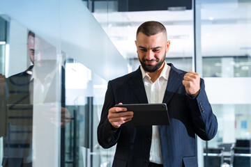Fototapeta premium Successful businessman winner with tablet computer made a good deal. Smiling man in business suit celebrates success by looking at the screen of a gadget.