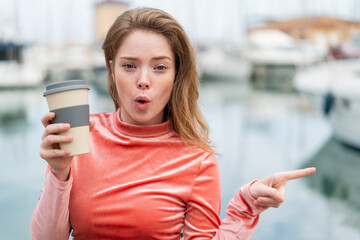 Young redhead woman holding a take away coffee at outdoors surprised and pointing side